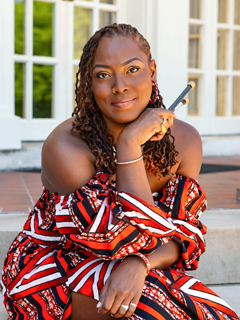 Photo of Dr. Josanne Francis seated outdoors on stirs of a building and holding a pair of steelband sticks.